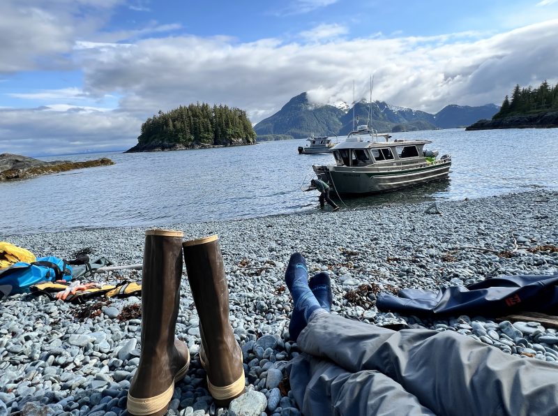 A person’s feet stretch out on a rocky beach with boots to the side and equipment around.