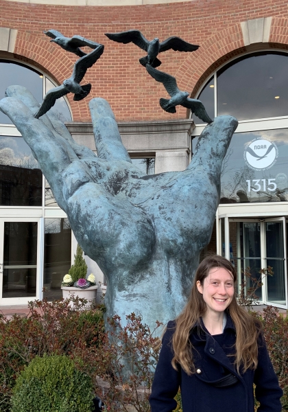A person stands in front of a hand shaped sculpture and smiles for a picture.