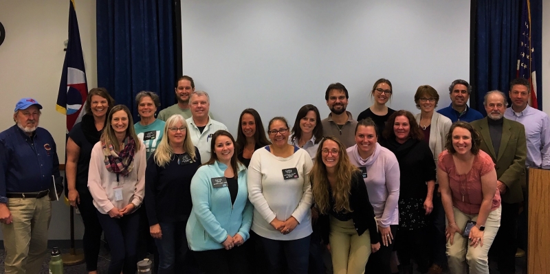 21 people stand in a row smiling for a picture. They are standing inside, in front of a blank wall.