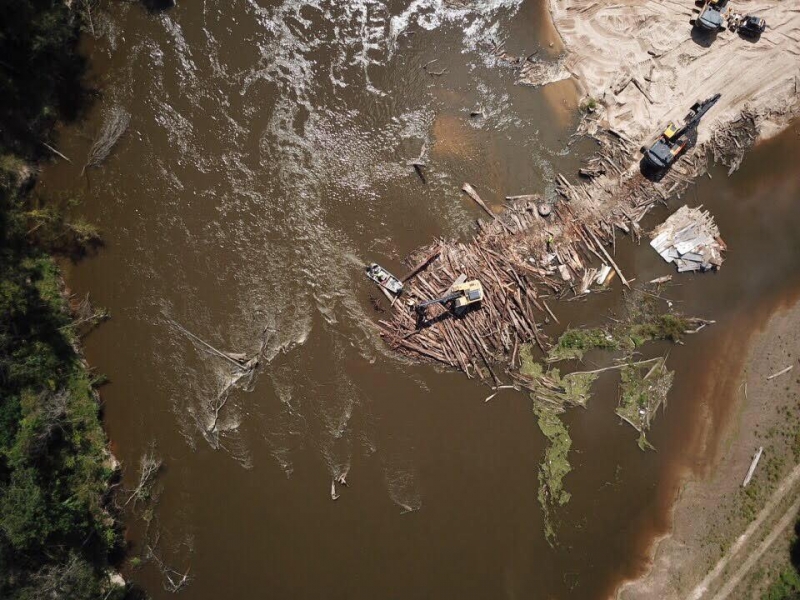 Several large construction excavators remove logs from a muddy river.
