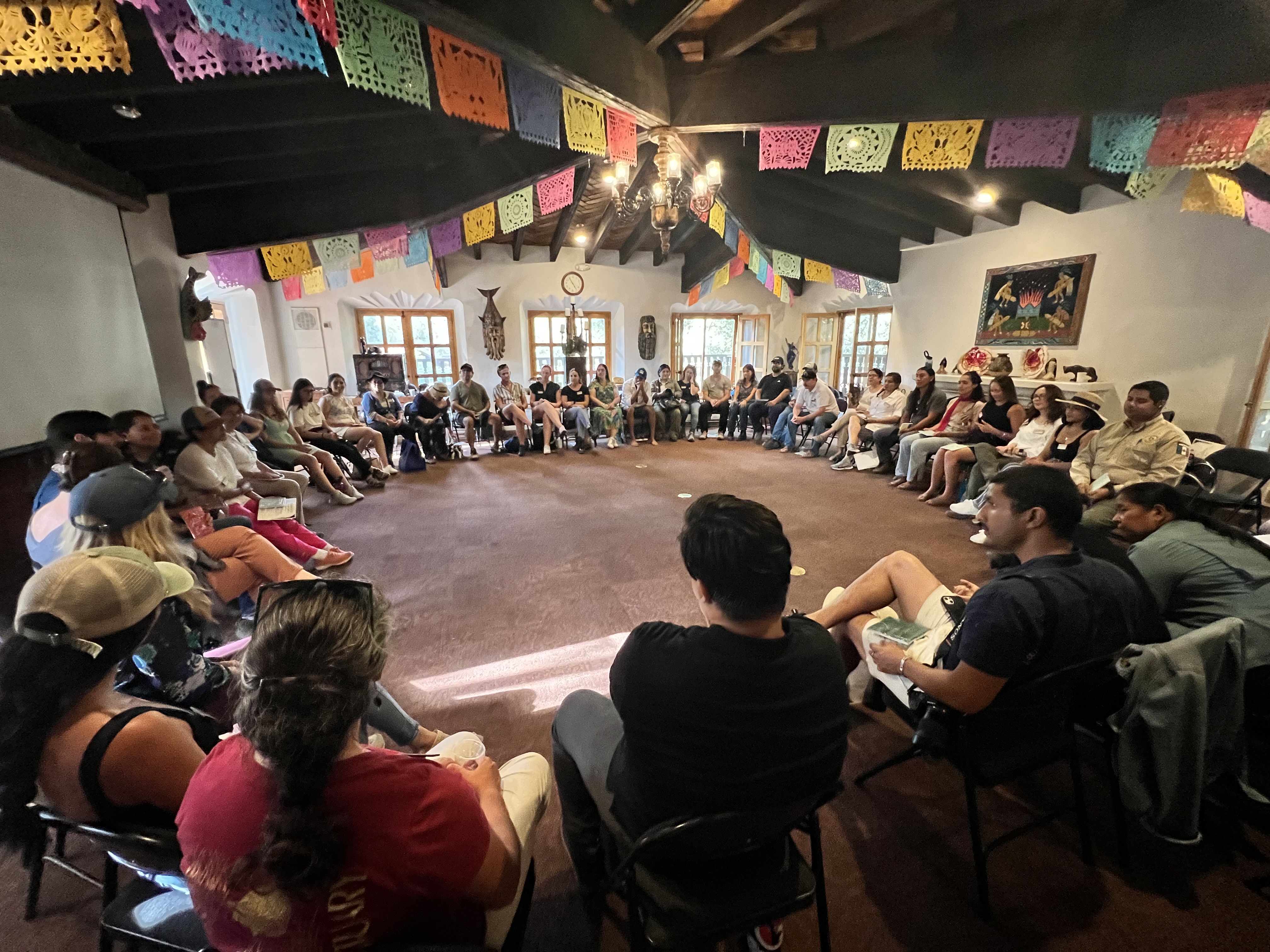 A group of people sit in a room with colorful flags hanging from the ceiling.