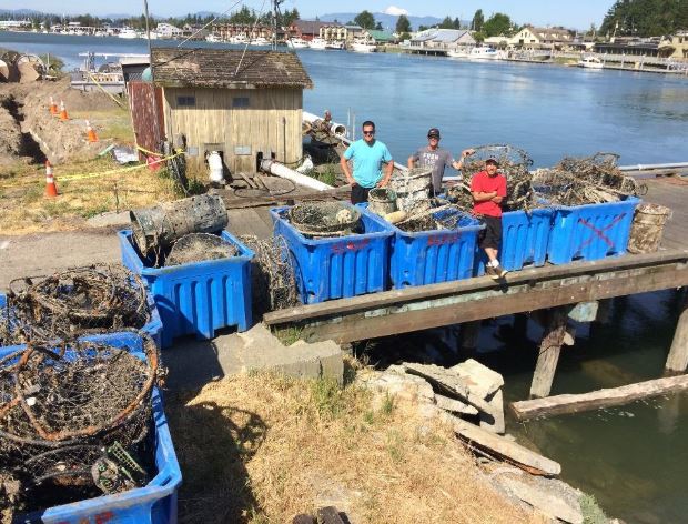 Three people stand on a pier that holds seven large, blue plastic bins that are completely filled with old crab pots that were pulled from the sea. 