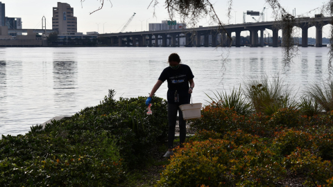 A student with gloves and a bucket picking up trash along a river shore.