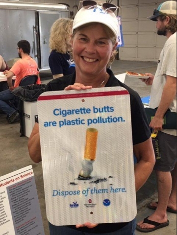 A person smiles while holding a sign that says "Cigarette butts are plastic pollution".