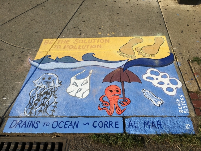 A painting over a sewer drain that reads "Drains to Ocean" and pictures human feet, jellyfish, octopus, plastic bag, and six pack rings.