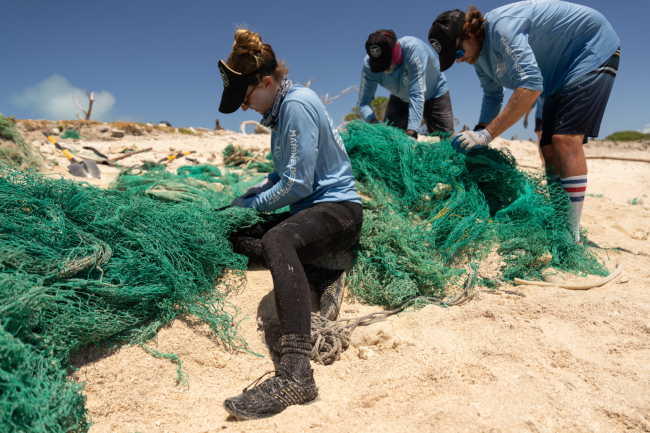  3 people cut up a large net on a beach.