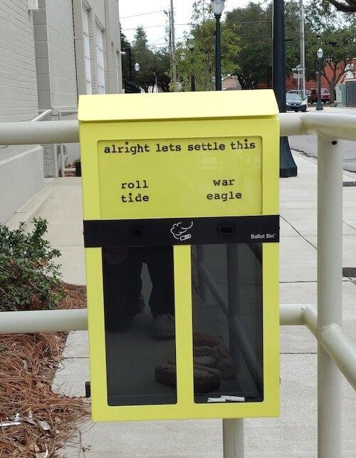 Yellow trash bin for cigarette butts split into two sides, ‘roll tide’ or ‘war eagle’.