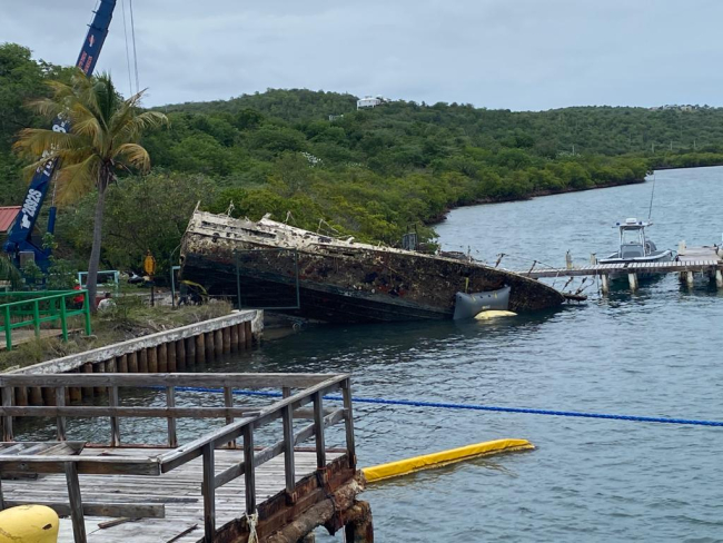 A derelict vessel grounded on its side on a shoreline near a dock.