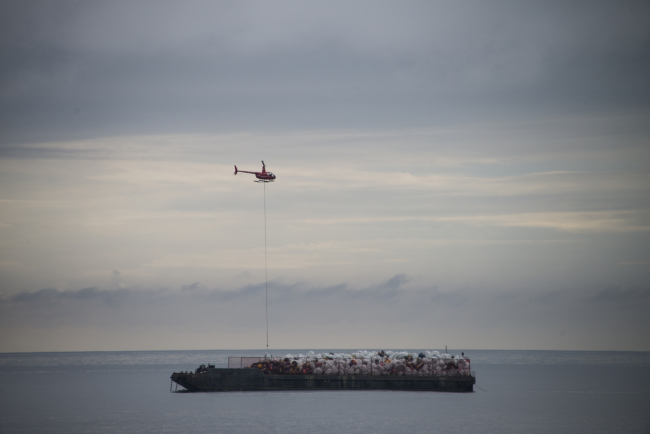 A helicopter hovering over a barge in the open water that is piled high with trash bags full of marine debris.
