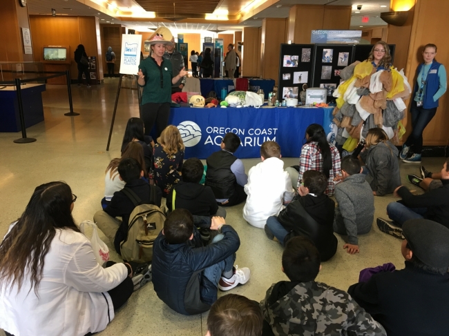 Three adults stand before a small audience who are all sitting on the ground. They are speaking about marine debris.