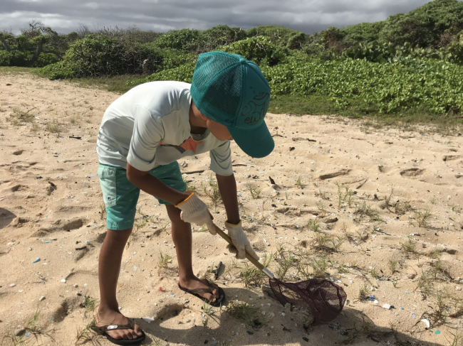 A child uses a small handheld net to sift trash from the sand on a beach.