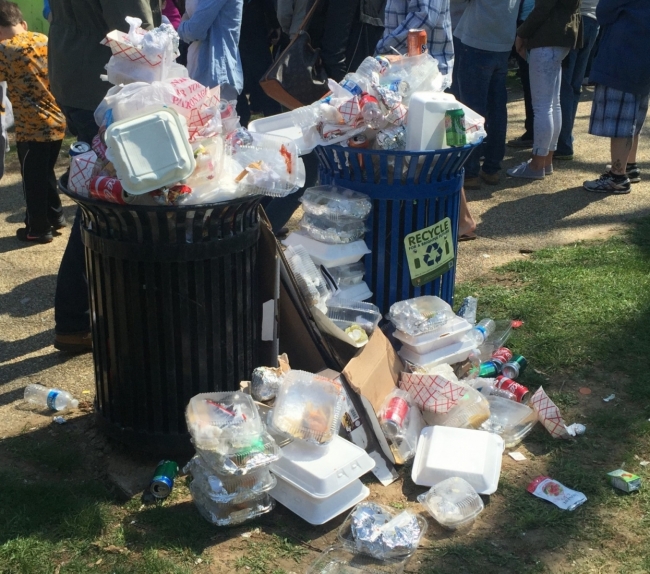 Two overflowing garbage cans with lots of garbage piled up on the ground by the cans.