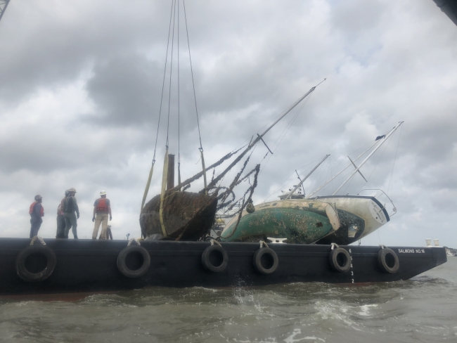Four people stand on a barge that holds three derelict sailboats.