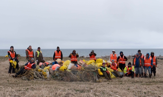 16 people stand around a large pile of collected marine debris and smile for a picture.