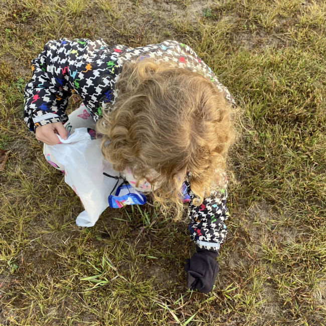 A child with a trash bag picks up litter from the grass.