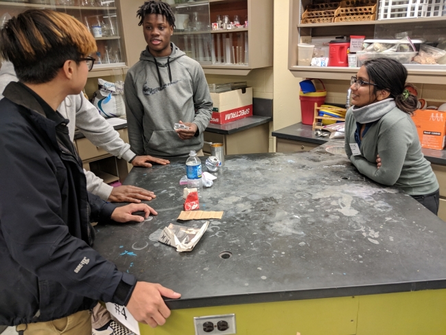 Students stand around a table with a plastic bottle and chip bags.
