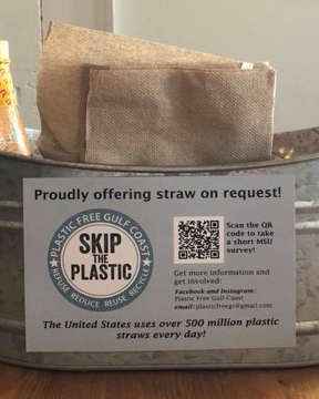 A metal tin sits on a wooden table and holds hot sauce and napkins. There is a sign on the tin that reads “Proudly offering straw on request! Skip the plastic.”