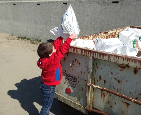 A child putting a bag of trash into a dumpster.