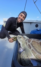 A person in a wetsuit sitting on the deck of a boat smiling and posing next to a green sea turtle resting in a container.