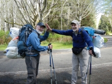 Two people “high five” each other while wearing very heavy backpacks full of collected marine debris. They also have full, plastic garbage bags attached to the backpacks. 