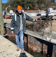 A person wearing a jacket and an orange beanie stands on a dock in the sun holding two retrieved crab trap debris items.