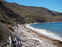 People appear small and spread out on a wide stretch of pebbled beach on a sunny day. 
