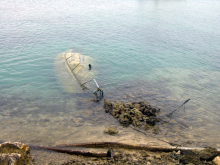 A sunken vessel that is completely submerged in clear water with its mast tangled in a rocky shoreline.