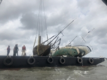 Four people stand on a barge that holds three derelict sailboats.