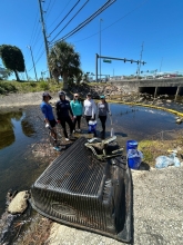 A group of people stand in shallow water in a concrete stream basin near large debris removed from a litter-trapping net. 